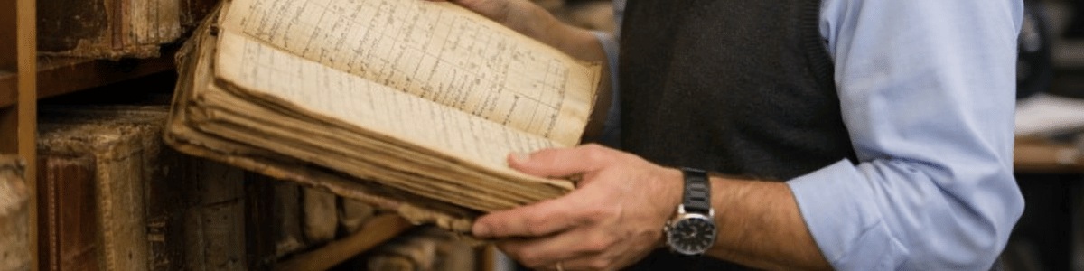Funcionário de cartório consulta cuidadosamente um antigo livro de registros em uma estante repleta de volumes antigos, em um ambiente institucional com iluminação suave e natural.