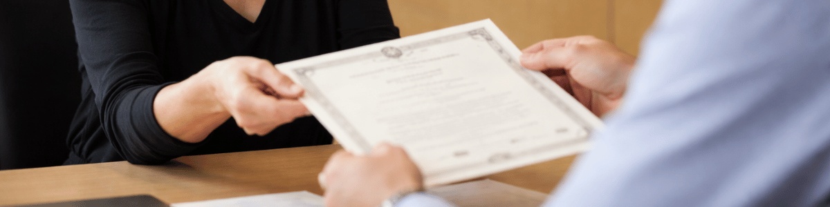 como averbar viuvez na certidão de casamento Uma mulher viúva, com expressão séria e contida, recebe uma certidão das mãos de um atendente em um cartório. O ambiente é interno, bem iluminado, com aparência institucional, e a cena transmite um momento delicado e formal, com foco nas mãos e no documento sendo entregue.