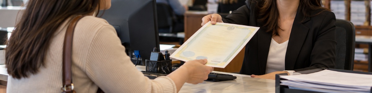 Uma pessoa recebe uma certidão das mãos de uma atendente em um cartório, em um ambiente organizado, profissional e acolhedor